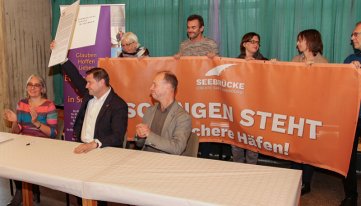 Die Urkunde wird hochgehalten vor dem orangen Banner der Seebrücke.